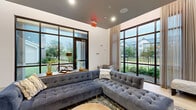This thoughtfully designed single-floor amenity space combines contemporary style with a welcoming, social atmosphere. The lounge area serves as the centerpiece, featuring tufted velvet sofas in rich navy and steel blue tones arranged on patterned area rugs atop light wood-style flooring. Soaring ceilings amplify the sense of openness, while expansive floor-to-ceiling windows framed in dark metal flood the interior with natural light and offer generous views of the surrounding greenery and outdoor landscaping. Sheer and solid drapery panels add an elegant, layered finish to the windows throughout.

A dedicated game room area provides a polished billiards table alongside wall-mounted cue storage, a large flat-screen television, and decorative framed artwork, creating an engaging space for recreation and entertainment. An adjacent kitchenette features white cabinetry with dark hardware, a gray quartz countertop, stainless steel appliances, and a striking gray subway tile backsplash that extends to the ceiling, with a wall-mounted television completing the space.

Throughout the lounge, curated décor including decorative sculptures, framed prints, and accent mirrors contribute to a refined yet approachable aesthetic. The seamless connection between seating zones, entertainment areas, and the kitchenette makes this space well-suited for gathering, relaxation, and community engagement.