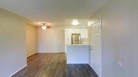 This single-floor residence offers a clean, modern aesthetic with an open-concept design that seamlessly connects the living area to the kitchen. The living space is finished with rich, dark hardwood-style flooring that extends throughout, providing a warm and grounded ambiance. A ceiling fan is centrally mounted, offering both comfort and a functional design element.

The kitchen is positioned at the heart of the space, defined by a peninsula-style breakfast bar that serves as a natural divider between the cooking and living areas. White cabinetry lines the upper section of the kitchen, providing ample storage while contributing to a bright, airy feel. A stainless steel refrigerator anchors the kitchen, complementing the contemporary cabinetry and light-toned countertops.

The walls are finished in a neutral, light tone that enhances the sense of openness and allows natural light to reflect throughout the space. A paneled entry door is visible, adding a classic architectural touch. The overall layout is practical and well-proportioned, making efficient use of the available space while maintaining a comfortable, livable atmosphere suitable for everyday use.