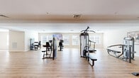 Located at 6039 Seminole Trail in Barboursville, Virginia, this fitness center offers a generously sized, single-floor workout facility designed to accommodate a wide range of exercise needs. The space is finished with warm-toned hardwood-style flooring throughout, contributing to a clean, welcoming atmosphere that encourages activity.

The main workout area is thoughtfully organized to maximize usability. A robust selection of strength training machines is distributed across the floor, including cable and multi-function weight machines, adjustable benches, and a well-stocked dumbbell rack with color-coded free weights. A dedicated stretching and functional training zone features stability balls, medicine balls, and a fold-out mat, providing ample room for dynamic warm-ups and cooldowns.

Cardio equipment, including stationary bikes and other aerobic machines, lines portions of the room, each positioned near the large windows that line the walls. These windows invite abundant natural light and provide outdoor views, creating an airy, open ambiance. Wall-mounted flat-screen televisions are strategically placed for entertainment during workouts.

The interior features neutral beige walls, recessed and flush-mount lighting, and a tray ceiling detail that adds subtle architectural definition. A mirrored wall expands the sense of space visually, while glass-panel entry doors provide a bright, accessible entryway. The overall design is functional, modern, and well-maintained.