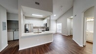 This single-story home is defined by its vaulted ceilings, which lend an airy, expansive quality to the main living areas. Dark hardwood-style flooring runs throughout the common spaces, contrasting cleanly with the neutral gray walls and bright white trim. An arched hallway entry adds an architectural detail that transitions smoothly into the open living area.

The kitchen is a central highlight, featuring white shaker-style cabinetry, light quartz countertops, a gray tile backsplash, and stainless steel appliances including a refrigerator, microwave, and range. A peninsula with a built-in sink and breakfast bar overhang connects the kitchen to the living space, making it well-suited for everyday use and entertaining.

The bedrooms are carpeted for comfort and include ceiling fans and windows that allow natural light. One bedroom offers direct access to the bathroom and additional closet space.

The bathroom is finished with a marble-patterned tile tub surround, an extended white vanity with ample storage, a wide mirror, and dark hardwood-style flooring. A secondary utility area with an electrical panel and closet space provides additional practical storage. Overall, the home presents a cohesive, contemporary aesthetic throughout.