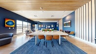 This single-floor community lounge is a thoughtfully designed multipurpose space that balances function and aesthetic appeal. The interior is immediately defined by its striking navy blue accent walls, which create a bold, cohesive backdrop throughout. Light hardwood flooring runs continuously across the space, complementing the warm-toned wood slat ceiling panels that add architectural texture and visual warmth.

The space is divided into two primary zones. A central dining and gathering area features a large, light-toned rectangular table surrounded by cognac leather chairs set atop a deep blue area rug, ideal for group meetings or shared meals. A vertical wood slat partition elegantly separates this zone from the adjacent lounge area.

The lounge zone is furnished with a leather sofa, rounded upholstered armchairs, and a wall-mounted flat-screen television, creating a relaxed, communal atmosphere. A walnut-toned media console anchors the seating area, while an eclectic collection of colorful artwork decorates the walls throughout.

Large black-framed windows and glass doors flood the space with natural light and offer outward views of the surrounding streetscape. Potted plants are distributed throughout, adding organic life to the vibrant, contemporary interior.
