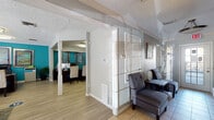 This single-story professional office space is thoughtfully designed to balance functionality with an inviting aesthetic. The layout flows naturally from a front reception and waiting area through to dedicated workspaces, offering a well-organized environment suited for client-facing operations.

The entrance and waiting area features light ceramic tile flooring, textured walls with a subtle striped pattern, and comfortable gray upholstered seating arranged around a small side table. A pendant light fixture and a glass-paneled front door allow natural light to filter into the space, creating an open and welcoming first impression. Crown molding and a textured ceiling add architectural character throughout.

The reception workspace is defined by a dark wood desk equipped with dual monitors, set against a bold teal accent wall adorned with framed decorative artwork. Flush-mounted ceiling fixtures provide consistent, even lighting, while light wood-style flooring transitions seamlessly from the front areas into the workspaces.

A private office features a matching dark wood L-shaped desk, a leather executive chair, and additional guest seating. Framed credentials and artwork decorate the walls, complementing the teal color scheme.

A separate conference or meeting room offers a rectangular wood table surrounded by yellow upholstered chairs, set atop a patterned area rug over wood-style flooring, providing an ideal space for collaborative discussions.