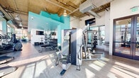 This fitness center in San Diego, CA, occupies a generously sized single-floor layout designed to accommodate a comprehensive range of workout activities. The space is defined by its industrial-modern architectural style, featuring exposed concrete ceilings with visible ductwork, large ceiling fans, and light wood-style flooring throughout the main workout areas.

Expansive floor-to-ceiling windows line multiple walls, flooding the space with abundant natural light and offering open views of the surrounding San Diego cityscape, including palm trees and neighboring buildings. The bright, airy atmosphere is complemented by bold teal accent walls and ceilings in select zones, adding a vibrant visual contrast to the industrial elements overhead.

The main workout floor is comprehensively equipped with a wide selection of strength machines, cable systems, free weights, medicine balls, elliptical trainers, treadmills, stair climbers, rowing machines, and spinning bikes. Mirrored walls in the free weights area enhance the sense of space. A dedicated free-weights and functional training zone features rubberized flooring for safety and versatility.

A standout feature is the indoor golf simulator room, outfitted with a full-screen projection system, artificial turf hitting surface, and a dark navy geometric-patterned accent wall, offering a unique recreational amenity within the facility.