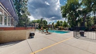 Located at 7520 W Amherst Ave in Denver, Colorado, this outdoor recreational amenity area serves as a central gathering space within the community. At its heart is a generously sized swimming pool with clear, blue water and lane markers, designed to accommodate both leisure swimming and lap swimming. The pool is enclosed by a secure perimeter fence, ensuring safety while maintaining an open and welcoming atmosphere.

The surrounding pool deck is finished in light concrete, providing ample space for lounging. Multiple sling-style lounge chairs and seating areas are arranged throughout the deck, offering residents convenient spots to relax and soak up the Colorado sun. A shaded pergola canopy structure on one side of the deck provides a comfortable retreat from direct sunlight, furnished with additional seating and tables for outdoor dining or socializing. Freestanding market umbrellas further supplement the shaded seating options across the deck.

The entire area is framed by an impressive collection of mature trees, including towering pines and leafy deciduous varieties, which lend the space a lush, park-like setting and provide natural shade. The backdrop includes the surrounding residential buildings, which feature a warm, earthy color palette with blue accent trim. A clubhouse-style structure with a covered porch overlooks the pool, adding a functional and architectural focal point to the space. The overall ambiance is relaxed, inviting, and well-suited for outdoor recreation.