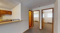 This single-floor unit offers a practical and well-organized layout across several distinct areas. The living room is a comfortable, carpeted space featuring a ceiling fan with integrated lighting, a wall-mounted HVAC unit, and a window fitted with blinds that allows natural light to filter in. The room transitions seamlessly into the entryway, which features hardwood-style flooring and a front door providing direct exterior access.

The kitchen is positioned adjacent to the living area and is accessible via a pass-through counter with an overhead opening that maintains visual connectivity between spaces. The kitchen includes warm-toned wood cabinetry, a white refrigerator, and a sink set beneath upper cabinets — a compact but efficiently arranged workspace.

A hallway connects the living area to the private rooms, featuring a built-in shelf and access to multiple doors. The bedrooms are carpeted and finished with neutral walls, textured ceilings, and windows with blinds. One bedroom includes a set of sliding wood-panel closet doors offering generous storage. Baseboard heaters are present in the bedrooms, ensuring consistent warmth. Throughout the unit, warm wood-toned door and window trim adds cohesion, while the textured ceilings are a consistent architectural detail across all rooms.