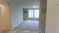 This single-floor unit offers a clean, functional layout with neutral-toned interiors throughout. The main living area features wall-to-wall carpeting in a light beige tone, smooth walls painted in a soft, warm neutral, and standard-height ceilings with flush-mount lighting fixtures. A transition from tile flooring near the entry to carpet in the main living space delineates the layout naturally.

Large windows fitted with vertical blinds allow generous amounts of natural light to fill the rooms, offering views of the surrounding outdoor greenery. Baseboard heating units are positioned beneath the windows, providing efficient climate control across the space. A wall-mounted air conditioning unit in the living area offers supplemental cooling during warmer months.

The walls are clean and unadorned, providing a blank canvas for personalization. White baseboards run consistently throughout, framing the carpeted floors neatly. An electrical panel is visibly mounted on one wall, conveniently accessible. The overall ambiance is modest and practical, with a neutral palette and straightforward finishes that offer a versatile foundation for a variety of interior design preferences.