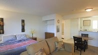 This single-floor space features an open-concept layout that seamlessly integrates the sleeping and living areas into one cohesive environment. The room is finished with smooth, light-colored walls and a clean white ceiling that helps reflect light and enhance the sense of openness. A sofa with accent pillows anchors the living area, positioned adjacent to the sleeping zone, which is furnished with a bed dressed in a striped coverlet. A wooden dresser with multiple drawers provides practical storage, while a built-in shelving nook in the wall offers additional display or storage space. A small nightstand with a vase of yellow florals adds a touch of warmth to the space. A decorative clock mounted on the wall adds character alongside framed wall art. The layout extends toward what appears to be a separate area — likely a bathroom or hallway — accessible through an open doorway, suggesting a degree of functional separation within the unit. A small dining or accent table with a dark chair is partially visible, rounding out the space's versatility. Overall, the space is practical and efficiently arranged.