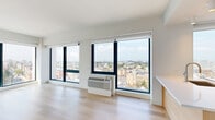 Located in Jersey City, NJ, this thoughtfully designed one-bedroom, one-bathroom residence offers a contemporary living experience with an abundance of natural light and sweeping urban views. Light hardwood flooring flows throughout, complementing the clean, modern aesthetic of the white-painted walls and minimalist finishes.

The open living room is a standout, featuring expansive floor-to-ceiling windows on multiple walls that frame panoramic views of the Jersey City skyline and surrounding neighborhoods, filling the space with natural light throughout the day. The kitchen is equally impressive, boasting large-format windows with city views, light wood cabinetry, stainless steel appliances including a French door refrigerator, microwave, and range, creating a bright and functional cooking space.

The generously sized bedroom continues the floor-to-ceiling window theme, offering additional sweeping city views and a dedicated climate control unit. A closet provides ample storage. The bathroom is finished with large-format neutral tiles, a soaking tub with shower, a sleek navy vanity with white countertop, and a lighted mirror cabinet. A dedicated laundry area and hallway round out this well-appointed home.