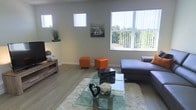 This single-floor space offers a welcoming and well-lit living area defined by clean lines and a contemporary aesthetic. The living room is anchored by large vertical-blind windows that invite generous amounts of natural light while framing lush outdoor greenery, creating a refreshing indoor-outdoor connection. Light hardwood flooring runs throughout, lending warmth and continuity to the space.

The room is thoughtfully furnished with a spacious gray sectional sofa paired with vibrant orange cube ottomans that add a bold pop of color. A glass-top coffee table sits atop a plush white area rug, contributing to the room's layered, modern feel. Decorative accents, including a sailboat wall print and potted plants, add personality and life to the space.

A media console with a television is positioned along one wall, complemented by additional high windows that bring in supplemental light from multiple angles. The neutral gray walls serve as a versatile backdrop that highlights the carefully chosen furnishings and décor. The overall ambiance is bright, airy, and casually sophisticated, making it an inviting space for both relaxing and entertaining.