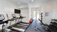 This fitness room offers a functional and well-organized workout environment designed to support a range of exercise routines. The space features crisp white walls and recessed lighting, creating a bright and clean atmosphere conducive to physical activity. Dark rubber flooring lines the room, providing durability and stability underfoot.

Cardio equipment is a focal point, with multiple TRUE-brand treadmills positioned along one wall, accompanied by an upright stationary bike. A wall-mounted flat-screen television is installed above the cardio machines, allowing users to stay entertained during their workouts. Supplementing the cardio area, the space includes stability and medicine balls for functional training, as well as a weight bench and a rack of dumbbells for strength exercises.

Natural light enters the room through a set of glass French doors at the far end, which also provide direct access to an outdoor area. An emergency exit sign is clearly visible, reflecting attention to safety compliance. The overall layout is efficient, making good use of the available space while maintaining an open, uncluttered feel suitable for a variety of fitness activities.