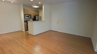 This 2-floor space features a bright, open-concept main level that seamlessly connects the kitchen and living area. The living space is finished with light hardwood flooring that extends throughout, lending warmth and continuity to the layout. Neutral white walls create a clean, versatile backdrop, while recessed lighting ensures consistent illumination across the space.

The kitchen is positioned behind a half-wall peninsula, which serves as both a functional workspace and a natural divider between the cooking and living zones. It is equipped with stainless steel appliances, including a refrigerator, stove, and over-the-range microwave, alongside medium-toned wood cabinetry offering ample storage. A skylight above the kitchen adds a natural light source to the area.

A paneled white door on the main level provides access to the exterior or an adjacent space. The open floor plan promotes a sense of spaciousness and ease of movement between areas. The overall ambiance is clean and functional, with a straightforward design that offers a solid foundation for personalization across both floors of the space.