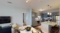 Located at 4001 Reedybrook Xing in Apex, North Carolina, this single-floor residence offers a thoughtfully designed living space with modern finishes and an inviting open-concept layout. The kitchen is a standout feature, showcasing sleek gray cabinetry, light granite countertops, a classic white subway tile backsplash, and stainless steel appliances, including a dishwasher. A kitchen island provides additional prep space and serves as a natural transition into the dining area. Track lighting illuminates both the kitchen and adjoining spaces, lending a bright and functional ambiance throughout. The dining area comfortably accommodates a full table and chairs, making it well-suited for everyday meals and entertaining alike. Warm hardwood-style flooring grounds the main living and kitchen areas, while a hallway visible from the main space leads to additional rooms finished with plush carpeting. The interior palette features soft, neutral wall tones that complement the contemporary design elements. The overall atmosphere is clean, modern, and move-in ready, offering a comfortable and practical layout for everyday living in the Apex, NC area.