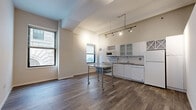 This thoughtfully laid-out one-bedroom, one-bathroom residence spans a single floor and makes excellent use of its compact footprint. The open living area is defined by rich dark hardwood flooring, impressively tall ceilings, and large windows that frame views of the surrounding urban streetscape, flooding the space with natural light. Track lighting overhead adds a modern, industrial accent to the airy atmosphere.

The kitchen is seamlessly integrated into the main living space and features white cabinetry with glass-front upper panels, a subway tile backsplash, granite-style countertops, a stainless steel sink, a dishwasher, and a full-size refrigerator. A distinctive built-in wine rack adds a stylish, functional touch to the cabinetry ensemble.

The bedroom offers a comfortable and quiet retreat with soft carpeted flooring, neutral-toned walls, and generous ceiling height. A doorway leads directly into the bathroom, providing convenient access.

The bathroom is finished with large-format diagonal-patterned tile flooring, a full soaking tub with a tiled surround accented by a classic black-and-white border, a sleek white vanity with ample drawer storage, and chrome fixtures throughout. Clean lines and a light color palette give the bathroom a fresh, polished feel. Overall, this residence combines urban character with practical, modern living essentials.