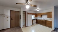 This single-floor space centers around a practical, open kitchen and living area layout. The kitchen is equipped with white appliances, including a gas range with oven and a full-sized refrigerator, complemented by a double-basin sink set beneath upper and lower cabinetry finished in warm medium-toned wood. The L-shaped counter configuration provides generous workspace, while the light-colored vinyl flooring adds brightness to the room.

A ceiling fan with a built-in light fixture serves as the central overhead lighting source, providing both illumination and air circulation throughout the combined space. An additional flush-mount ceiling light near the kitchen area ensures the workspace remains well-lit.

Adjacent to the kitchen, an open doorway reveals a built-in storage closet fitted with multiple shelves, offering practical organizational space. A solid wood entry door is positioned nearby, providing access to the unit.

The transition from the kitchen's vinyl flooring to carpeted flooring delineates the living area, adding warmth and comfort underfoot. The overall layout is straightforward and functional, designed to maximize utility within a compact, single-level footprint.