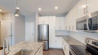 This single-floor unit in Chapel Hill, NC offers a thoughtfully designed layout with contemporary finishes throughout. The kitchen is a standout feature, showcasing white shaker-style cabinetry extending to the ceiling, granite countertops, a subway tile backsplash, and a full suite of stainless steel appliances including a refrigerator, electric range, and built-in microwave. Dark hardwood-style flooring anchors the kitchen and flows into the open living area, which is brightened by multiple windows, a ceiling fan, and glass doors leading to an outdoor space. The bedrooms feature neutral carpet flooring and windows that allow natural light to filter in, creating a calm atmosphere. A walk-in closet with multi-level wire shelving provides generous storage. The bathrooms are finished with granite-topped white vanities and wood-look tile flooring. One bathroom includes a tub-shower combination with white tile surround, while the second features an elongated vanity layout. Soft gray wall tones carry throughout the unit, contributing to a clean, modern, and cohesive aesthetic.