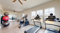 Located at 109 Alicia Way in LaGrange, Georgia, this fitness center offers a well-appointed, single-level workout space designed to support a comprehensive exercise routine. The room features a light-toned, durable floor finish that provides a clean and functional surface throughout the space.

Natural light fills the room through multiple large, multi-pane windows along the walls, offering views of the surrounding neighborhood and contributing to an open, energizing atmosphere. Ceiling fans are installed overhead to maintain airflow and comfort during workouts.

The space is equipped with a variety of cardio machines, including treadmills and elliptical trainers, positioned to take advantage of the natural light from the windows. A dedicated free weights area features a full rack of dumbbells and a weight bench, accommodating strength training needs. Exercise balls and additional fitness accessories are also present, supporting flexibility and cross-training activities.

A large wall mirror spans one side of the room, visually expanding the space and allowing users to monitor their form during exercise. Built-in shelving along one wall provides convenient storage. The overall design is clean and minimalist, with neutral white walls and recessed lighting complementing the bright, motivating environment of this practical and fully equipped fitness facility.