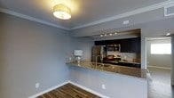 This single-floor residence showcases a thoughtfully designed open-concept layout that seamlessly connects the kitchen and living areas. The kitchen is a standout feature, equipped with stainless steel appliances including a refrigerator, microwave, and gas range. Dark espresso cabinetry provides bold contrast against the granite countertop peninsula, which doubles as a breakfast bar and serves as a natural dividing element between the kitchen and the adjoining living space. A decorative stone tile backsplash adds texture and visual interest behind the cooking area, while under-cabinet track lighting ensures practical illumination for meal preparation. The living area is finished with wood-look tile flooring in a warm tone, offering durability with an elegant aesthetic. Walls painted in a cool slate gray create a calm, cohesive atmosphere throughout the space. Crown molding runs along the ceiling perimeter, lending a refined, polished character to the interior. A flush-mount ceiling light fixture with a fabric shade provides warm ambient lighting. White baseboards provide crisp contrast against both the flooring and walls, completing the clean, modern look of this well-appointed residence.