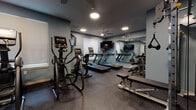 Located at 533 Pinney St in Madison, WI, this fitness center is a well-appointed, single-level facility designed to support a comprehensive workout experience. The space is finished with durable rubber flooring in a speckled dark tone, providing both safety and comfort underfoot throughout the entire area.

The cardio section is prominently featured, housing multiple treadmills positioned along one wall, each paired with wall-mounted flat-screen televisions for entertainment during workouts. Flanking the treadmills are several elliptical and cross-trainer machines, offering a range of low-impact cardio options. Ceiling fans are installed overhead to maintain airflow and comfort throughout exercise sessions, complemented by recessed lighting that keeps the space well-lit.

A large floor-to-ceiling mirror spans one wall, visually expanding the room and allowing users to monitor their form during training. The strength and functional training area includes a tiered dumbbell rack with a full range of free weights, a weight bench, and a colorful assortment of stability balls, medicine balls, and resistance equipment neatly organized on a dedicated storage rack. Natural light filters in through blinded windows, and the muted blue-gray walls contribute to a focused, energizing atmosphere throughout the facility.