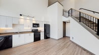 This contemporary two-story loft-style unit showcases an open, airy design defined by soaring double-height ceilings on the main floor. The open-concept living and kitchen area is anchored by a sleek galley-style kitchen featuring white flat-front cabinetry with under-cabinet lighting, warm brown countertops, a stainless steel undermount sink, and a full suite of black appliances including a range, microwave, dishwasher, and refrigerator. Light wood-tone luxury vinyl flooring runs throughout the main level, complementing the crisp white walls and modern aesthetic. A striking staircase with a black metal railing leads to the upper floor, where the bedroom offers generous floor-to-ceiling windows that frame views of the surrounding urban streetscape and fill the room with natural light. The bedroom is accompanied by a walk-in closet with wire shelving. The upper level also includes a bathroom featuring a soaking tub with full tile surround. A half bathroom is conveniently located on the main floor, finished with a large vanity mirror and dark wood-tone shelving. Throughout the unit, dark door and window trim provides a clean contrast to the neutral palette, contributing to a polished, contemporary look.