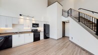 This contemporary two-story loft-style unit showcases an open, airy design defined by soaring double-height ceilings on the main floor. The open-concept living and kitchen area is anchored by a sleek galley-style kitchen featuring white flat-front cabinetry with under-cabinet lighting, warm brown countertops, a stainless steel undermount sink, and a full suite of black appliances including a range, microwave, dishwasher, and refrigerator. Light wood-tone luxury vinyl flooring runs throughout the main level, complementing the crisp white walls and modern aesthetic. A striking staircase with a black metal railing leads to the upper floor, where the bedroom offers generous floor-to-ceiling windows that frame views of the surrounding urban streetscape and fill the room with natural light. The bedroom is accompanied by a walk-in closet with wire shelving. The upper level also includes a bathroom featuring a soaking tub with full tile surround. A half bathroom is conveniently located on the main floor, finished with a large vanity mirror and dark wood-tone shelving. Throughout the unit, dark door and window trim provides a clean contrast to the neutral palette, contributing to a polished, contemporary look.