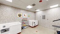 Located at 5801 Sanderson St NW in Huntsville, Alabama, this purpose-built pet grooming room offers a clean, well-equipped single-room space designed specifically for dog grooming and bathing. The room's most distinctive architectural feature is its floor-to-ceiling terrazzo-patterned wall paneling, which lends a durable, easy-to-clean surface throughout the space while giving it a bright, speckled aesthetic. A suspended drop ceiling hosts multiple recessed flat lighting panels that ensure consistent, even illumination across the entire room. The polished concrete floor adds to the practical, maintenance-friendly character of the space. The room is outfitted with a large, white walk-in dog bathing tub featuring a step-entry design for easy pet access, a dedicated dog grooming table equipped with a professional-grade stand dryer and arm attachment, a utility sink station along one wall, and a multi-tier metal shelving unit stocked with grooming supplies and treats. Cheerful, dog-themed framed artwork adorns the walls, adding a welcoming and playful touch to the functional environment. A wall-mounted information panel provides guidance for users of the space. The room is accessed through a solid white door and includes an HVAC vent and fire suppression system, reflecting attention to safety and comfort. Overall, the space presents as a clean, organized, and fully equipped self-serve or professional pet grooming facility.