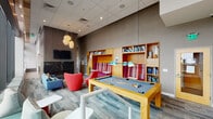 This dynamic communal amenity space is designed to foster relaxation, socializing, and recreation in equal measure. The layout flows across a generously proportioned open floor plan with soaring ceilings that lend a sense of grandeur throughout.

At the heart of the space sits a full-size pool table with pendant lighting overhead, framed by built-in warm wood bookshelves and vibrant plaid booth-style seating. A dedicated lounge area features bold red accent chairs, a soft blue sofa, and a geometric coffee table, all anchored near a dramatic floor-to-ceiling black marble fireplace with vertical wood paneling above — creating an inviting focal point.

Floor-to-ceiling windows flood the space with natural light and open to breathtaking panoramic city skyline views, with access to an outdoor terrace complete with lounge chairs. The mix of patterned carpeting and hard flooring adds textural contrast throughout.

A separate wet bar area offers a long grey countertop with built-in stainless steel appliances, a wall-mounted television, and a row of mint green barstools — ideal for casual entertaining. Colorful artwork and carefully curated pendant lighting complete the modern, eclectic aesthetic, making this a well-appointed space for both leisure and gathering.