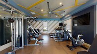 This fitness center offers a comprehensive workout environment designed with both functionality and visual appeal in mind. The space features a bold, contemporary aesthetic defined by a striking ceiling with yellow geometric accent beams set against a blue-gray backdrop, complemented by recessed lighting and a large industrial-style ceiling fan that enhances air circulation throughout the room.

The flooring consists of durable rubberized material suited for high-traffic fitness use, providing cushioning and traction across the entire area. Equipment is well-distributed throughout the space and includes treadmills, elliptical trainers, a stationary bike, a multi-station cable and strength machine, and a dedicated free weight area with a tiered dumbbell rack and adjustable bench. Multiple wall-mounted flat-screen televisions are positioned throughout the room for viewing during workouts.

Full-length mirrors along one wall visually expand the space while allowing users to monitor their form. Natural light enters through several windows fitted with blinds, and a set of glass double doors opens directly to an outdoor area with views of a pool and surrounding landscaping, creating a bright and airy atmosphere. The diagonal stripe wall mural adds a dynamic graphic element to the overall design.