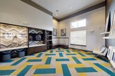 Located at 23400 Kingsland Blvd in Houston (Katy), TX, this stylish single-room lounge space offers a thoughtfully designed interior that blends contemporary aesthetics with functional comfort. The room is defined by its striking geometric-patterned carpet in teal, yellow, and neutral tones, which anchors the space and immediately draws the eye. Soaring ceilings with a two-tone treatment — crisp white above a bold dark gray band — lend an open, airy quality, while clerestory windows allow natural light to filter in from above.

A built-in dark wood media unit serves as a focal point along one wall, featuring a mounted flat-screen television, open shelving with chevron-patterned accents, and lower cabinetry for storage. Adjacent to the media wall, an upholstered built-in bench provides casual seating. A contrasting dark navy accent wall on the opposite side displays a curated gallery of eclectic framed artwork, complemented by floating shelves and wall-mounted digital panels. Additional seating is available throughout the room.

Glass French doors connect the lounge directly to a fitness center, enhancing the space's utility as a multi-purpose amenity area. The overall ambiance is energetic yet inviting, making it well-suited for social gatherings or relaxed lounging.