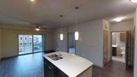 This single-floor space is designed with an open-concept layout that seamlessly connects the kitchen and living areas. The kitchen centers around a curved island with a white countertop and an undermount double sink, complemented by dark cabinetry below and a built-in dishwasher. Two cylindrical pendant lights hang above the island, providing focused task lighting while adding a modern aesthetic touch. Dark hardwood-style flooring runs throughout, creating a cohesive and contemporary look.

The living area extends naturally from the kitchen, featuring a ceiling fan for added comfort. A sliding glass door leads to a balcony, allowing natural light to filter in and offering views of the surrounding cityscape. The neutral gray walls throughout the space provide a clean, versatile backdrop. A doorway visible from the main living area connects to an adjacent room, carpeted in neutral tones, suggesting a bedroom or secondary living space. Built-in cabinetry near the entrance adds practical storage. The overall ambiance is modern, functional, and well-suited for everyday living.