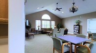 This inviting single-floor communal space is defined by its striking vaulted ceiling, which rises to a prominent peak and creates an impressive sense of openness and volume throughout the room. A large arched window anchors the far wall, flanked by additional rectangular windows that flood the interior with generous natural light and offer views of the surrounding greenery outside.

The space is thoughtfully arranged to serve multiple functions. A comfortable lounge area features plush sofas, upholstered armchairs, and side tables with warm lamp lighting, all positioned to encourage relaxed conversation and social gathering. Potted trees and decorative greenery add a natural, refreshing touch to the warm, neutral color palette of the walls and carpeted flooring.

Closer to the foreground, a game or activity table surrounded by cushioned chairs provides a dedicated area for organized group activities or informal meetings. A bookcase along one wall adds a homey, resourceful element to the space.

Ceiling fans and a chandelier-style light fixture complement the overhead recessed lighting, ensuring the room remains comfortable and well-lit throughout the day. Overall, the space strikes a balance between functionality and warmth, making it well-suited as a communal gathering area for leisure, socializing, and activities.