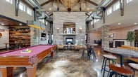 This single-story clubhouse blends rustic charm with industrial design across a thoughtfully arranged floor plan. The centerpiece is a grand great room with soaring vaulted ceilings supported by exposed wood trusses and visible ductwork, anchored by a floor-to-ceiling stacked stone fireplace. Wingback chairs arranged around the hearth create an inviting seating area, while a full-size pool table and high-top tables with industrial stools add to the social atmosphere. Clerestory windows allow natural light to pour into the space, complementing the rich, acid-stained concrete floors found throughout.

The kitchen area is a standout feature, boasting a corrugated metal island wrapped in dark granite countertops, a pressed tin ceiling with pendant lighting, stainless steel appliances including double wall ovens and a full-size refrigerator, open shelving, and a coffee station. A comfortable lounge area with a large-screen TV, leather seating, and natural light from large windows provides a relaxed gathering spot.

Additional amenities include dedicated game rooms with ping pong and air hockey tables, a well-lit corridor with water fountain access, and restroom facilities, making this a fully equipped communal facility.