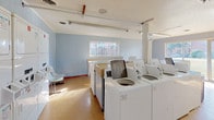 This single-floor laundry facility is designed to serve a communal setting efficiently. The space is characterized by light blue paneled walls, a textured white ceiling, and durable tile flooring throughout. Fluorescent lighting fixtures are installed along the ceiling, ensuring the room is well-lit at all times.

A central row of top-load washing machines and stacked dryer units lines the interior, providing ample capacity for multiple users. Each machine features individual control panels for straightforward operation. Adjacent to the machines, an extended built-in folding counter runs along one wall, offering generous workspace for sorting and folding laundry. Additional seating, including molded plastic chairs, is available for residents waiting during cycles.

Natural light enters through several windows, including a large sliding glass door that opens to a view of a grassy outdoor area. A bulletin board is mounted on the wall, serving as a space for notices and information. A trash receptacle is conveniently positioned near the machines. The overall layout is practical and spacious, allowing multiple users to move comfortably throughout the facility simultaneously.