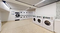 This well-appointed shared laundry facility offers a generously sized, single-floor layout designed to accommodate high-volume laundry needs with efficiency and comfort. The space is outfitted with a substantial number of commercial-grade front-loading washing machines and stacked dryer units arranged methodically along the perimeter walls, maximizing usable floor space while providing an impressive laundry capacity.

The interior design is clean and contemporary, featuring bright white walls and cabinetry that contribute to a fresh, hygienic atmosphere. Underfoot, speckled terrazzo flooring adds a durable and visually appealing finish that is easy to maintain. Overhead, a slatted ceiling with recessed linear LED light strips runs the length of the room, delivering consistent, even illumination throughout the facility.

Along one side of the room, a row of white countertop surfaces with integrated payment or control terminals provides convenient stations for users to manage their laundry. A textured decorative wall panel serves as a subtle design accent, breaking up the otherwise uniform white palette. A solid steel entry door with porthole windows provides access to the space, reinforcing the facility's functional, utilitarian character. Overall, the facility presents a highly organized, clean, and modern environment purpose-built for efficient communal laundry use.