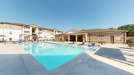 This beautifully designed outdoor amenity space centers around a large, resort-style swimming pool with crystal-clear blue water, surrounded by a generously sized concrete pool deck lined with lounge chairs — ideal for relaxing under open skies. The pool area is framed by multi-story residential buildings featuring balconies and neutral, warm-toned exteriors that create an inviting community atmosphere.

A standout feature of this space is the covered outdoor pavilion, anchored by distinctive natural stone columns that lend a rustic yet refined character to the setting. Beneath the covered structure, a ceiling fan keeps the space comfortable year-round. The pavilion includes a built-in bar counter with bar-height seating, perfect for casual entertaining. A cozy lounge area with plush sectional seating faces a wall-mounted flat-screen television above a sleek linear fireplace set into a natural stone surround, creating a warm and inviting focal point.

Adjacent to the pavilion, an open-air patio extends the outdoor living space, featuring additional seating areas with shade umbrellas and a built-in grill station under a pergola, making it well-suited for outdoor gatherings. The combination of the stone detailing, lush planters, and thoughtfully arranged furnishings gives this communal outdoor space a polished, resort-inspired aesthetic throughout.