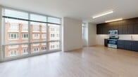 Located at 1939 Dorchester Ave in Boston's Dorchester neighborhood, this modern single-floor unit offers a thoughtfully designed layout with contemporary finishes throughout. The kitchen is a standout feature, outfitted with dark wood cabinetry extending to the ceiling for ample storage, sleek white countertops, a stainless steel refrigerator, range, microwave, and dishwasher, all complemented by light hardwood flooring. The open living area is filled with natural light, thanks to expansive floor-to-ceiling windows that frame views of the surrounding neighborhood and nearby brick architecture. The spacious layout flows seamlessly between the kitchen and living areas. Bedrooms are carpeted for comfort and equipped with individual wall-mounted mini-split HVAC units, offering personalized climate control. One bedroom features corner windows with views of the tree-lined streetscape below. A walk-in closet provides generous storage. The bathroom is finished in a clean, modern style with a white quartz vanity, an illuminated mirror, wood-toned cabinetry, and a full bathtub with accessible grab bars. White walls and abundant natural light create a bright, airy ambiance throughout the entire unit.
