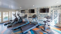 This fitness center is a thoughtfully designed workout space that combines functionality with energetic style. The room is immediately eye-catching thanks to its bold chevron-patterned flooring in shades of orange, teal, and grey — a dynamic design choice that sets an energetic tone throughout the space.

The cardio zone is well-appointed with multiple treadmills, an elliptical trainer, and a stationary bike, all positioned in front of wall-mounted flat-screen televisions for entertainment during workouts. A striking typographic accent wall lines the back of the room, featuring fitness-themed words such as "Endurance," "Strength," "Agility," and "Wellness" in varying fonts and sizes, reinforcing the motivational atmosphere.

The strength training area includes a multi-functional cable machine and a full dumbbell rack displayed in front of floor-to-ceiling mirrors, which enhance the sense of openness and allow users to monitor their form. Recessed ceiling lighting provides bright, even illumination throughout.

Natural light fills the space through large windows and a set of glass-paneled double doors at the entrance, which open to an outdoor courtyard area. The clean white walls and crisp finishes contribute to a fresh, modern aesthetic, making this fitness center both an inviting and practical space for a comprehensive workout routine.