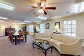 Located at 391 Old Griffin Rd in McDonough, Georgia, this single-floor community space offers a warm and functional layout designed for gathering and socializing. The main open area seamlessly combines a lounge and dining zone, carpeted throughout and bathed in natural light from multiple large windows fitted with blinds. The lounge is anchored by a curved sectional sofa, a glass-topped coffee table, and a ceiling fan with integrated lighting, creating a comfortable and inviting atmosphere. Framed artwork and table lamps add a refined touch to the warm, neutral-toned walls.

The dining section accommodates a long table with ample seating, making it well-suited for group meals or activities. A dedicated piano nook features an upright piano, a wooden bookcase, and additional seating, offering a quiet corner for leisure. A large mirror and black accent chairs line one wall near the entryway, providing a welcoming transition into the main space. Tall indoor plants are placed throughout, adding a natural, lively element to the interior.

The kitchen is a practical galley-style space with honey-toned wood cabinetry, laminate countertops, and a striking black-and-white checkered tile floor. It is equipped with a full-size refrigerator, electric range, microwave, and sink, supporting full catering or event needs. A utility corridor connects the kitchen to the main gathering area efficiently.