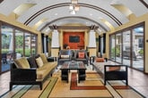 Located at 34875 Pourroy Rd in Winchester, CA, this single-floor living space offers a thoughtfully designed interior defined by striking architectural details and a warm, inviting ambiance. The centerpiece of the room is a dramatic barrel-vaulted ceiling adorned with dark accent arches and decorative brown trim, lending a sense of grandeur and vertical depth to the space. Skylights and clerestory windows positioned along the upper walls allow abundant natural light to filter in throughout the day, further enhancing the airy feel.

Large glass sliding doors and floor-to-ceiling windows line multiple walls, providing seamless transitions to outdoor patio areas and offering expansive views of the surrounding exterior. Dark wood-framed curtains frame the openings, adding a refined contrast to the warm cream and yellow wall tones.

The living area is furnished with a generous arrangement of sofas, accent chairs, and a glass-top coffee table set atop a bold geometric area rug in earth tones. A prominent white fireplace surround serves as a focal point along one wall, complemented by a wall-mounted television set against a rich terracotta-orange accent wall. A bar-height counter with seating provides additional functionality. Large-format tile flooring grounds the space with a clean, durable finish throughout.