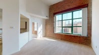 This distinctive urban loft-style space blends industrial character with comfortable modern living across a single floor. The expansive living area immediately impresses with soaring ceilings that amplify the sense of openness, soft carpeting underfoot, and dramatic exposed brick walls that serve as a defining architectural element throughout. Large green-framed windows flood the space with natural light while framing sweeping views of the surrounding cityscape, creating a striking connection between the interior and the urban environment outside.

The kitchen is well-appointed with ample white cabinetry accented by warm wood trim, generous counter space, tile flooring, and black appliances including a refrigerator, gas range, and built-in microwave. A pass-through opening connects the kitchen to the main living area, enhancing the open flow of the layout.

The bedrooms continue the loft aesthetic, featuring tall ceilings, plush carpeting, and exposed brick walls with city-facing windows that bring in abundant daylight. Closet space is provided with built-in shelving and hanging rods. Two full bathrooms are clean and functional, each featuring a tub-shower combination, vanity with mirror, and tile flooring. The overall ambiance of the space is bright, airy, and urban, making it a distinctive residence rich in architectural character.