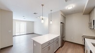 This single-floor unit offers a clean, contemporary design throughout, with a thoughtful layout that maximizes functionality and natural light. The open-concept kitchen serves as the heart of the space, anchored by a generously sized island topped with sleek white quartz countertops. Gray shaker-style cabinetry lines the walls, complemented by a white subway tile backsplash and a full suite of stainless steel appliances, including a French door refrigerator, electric range, microwave, and dishwasher. Floating metal shelves add an industrial-modern accent, while dual pendant lights with brass fixtures provide warm, focused illumination. The kitchen flows seamlessly into the open living area, which features light wood-tone flooring, neutral walls, and track lighting overhead. Large windows allow generous natural light to fill the living space. The bedrooms maintain the same light, airy palette with warm beige walls, wood-look flooring, and oversized windows fitted with roller shades for privacy and light control. The bathroom is finished with large-format tile on both the floors and tub surround, offering a clean and polished look. A rich wood-toned entry door adds a warm contrast to the otherwise neutral interior. Overall, the space presents a cohesive modern aesthetic that balances style with practicality.