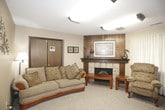This single-floor space is thoughtfully divided into two distinct functional areas, offering a blend of comfort and practicality. The lounge area anchors one end of the space and is highlighted by a tile-surround fireplace with a warm wood mantel, above which hangs a large framed artwork. Wood-paneled walls add a traditional, cozy character to this seating zone, which is furnished with a plush sofa and a patterned recliner arranged around a wooden coffee table. Warm, neutral carpet flows throughout, contributing to the relaxed ambiance.

Adjacent to the lounge is a more open, versatile meeting area. This section features light beige walls, a textured ceiling, and overhead fluorescent lighting that provides even, functional illumination. Multiple folding tables and chairs are arranged throughout, making the space adaptable for meetings, classes, or social gatherings. White curtain-covered windows are present along the walls, allowing diffused natural light to filter in while maintaining privacy. A wall-mounted heater and a side door provide additional utility. Framed artwork and subtle wall décor add modest character throughout the space.