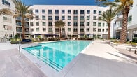 Located at 1881 N Rollins Rd in Burlingame, California, this outdoor amenity courtyard is a standout feature of a contemporary multi-story residential community. The centerpiece of the space is a generously sized resort-style swimming pool with sparkling blue water, surrounded by expansive light-toned pool decking finished in large-format tile. The pool area is framed by tall, mature palm trees that lend a California resort atmosphere, complemented by sun loungers and seating areas shaded by deep navy blue umbrellas. A secondary pool or spa area is also present, offering additional aquatic options for residents. The courtyard is enclosed on multiple sides by the building's modern white façade, characterized by large windows, glass-railed balconies, and clean geometric lines that flood the outdoor space with natural light. A welcoming passageway with a double-height ceiling and warm wood-accented architectural details serves as a formal entry point to the courtyard. Lush, curated landscaping — including raised planting beds, ornamental grasses, shrubs, and native-style plantings — adds greenery and texture throughout. Secured glass-panel entry gates provide access to pool areas while maintaining safety and privacy. The overall ambiance is bright, open, and resort-inspired, offering a refined outdoor retreat within an urban setting.
