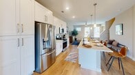 This three-story townhome in Charlotte's University Executive Park area offers a well-designed layout across three distinct floors, accommodating three bedrooms, four bathrooms, and an attached garage. The entry-level floor houses the garage and provides a functional base for the home. The main living floor serves as the heart of the residence, where an open-concept kitchen flows seamlessly into the living room. The kitchen is appointed with white shaker cabinetry, quartz countertops, stainless steel appliances including a French door refrigerator and range, a subway tile backsplash, and a center island with pendant lighting. The bright living room features hardwood-style flooring, a ceiling fan, and multiple windows that invite natural light throughout. French doors on the upper floor open to a private balcony, adding outdoor access to one of the bedrooms. Each of the three bedrooms includes a dedicated closet, while the bathrooms are finished with white vanities, quartz counters, and clean, neutral tones. Two staircases connect the floors, enhancing accessibility and flow throughout the home. The overall aesthetic is modern and cohesive, with warm neutral walls, consistent hardwood-style flooring, and contemporary fixtures across all levels.