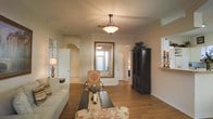 This single-story space exudes a traditional, elegant character anchored by warm hardwood flooring that extends throughout the main living area. The living room serves as the central gathering space, furnished with a neutral-toned sofa and ornately patterned accent chairs that complement the room's classic aesthetic. A dark, intricately carved armoire adds a stately focal point along one wall, while a large, gilded-frame mirror reflects natural light and amplifies the sense of space. A pendant light fixture with a frosted glass shade hangs from the ceiling, casting a soft, warm glow over the room. Crown molding frames the ceiling, adding architectural refinement, while arched doorways provide graceful transitions between rooms. A table lamp with a traditional base adds layered lighting near the seating area. Hallways branch off toward additional rooms, hinting at a thoughtfully connected floor plan. Wall sconces and windows allow natural light to filter in, enhancing the warm, inviting ambiance. Overall, the space balances classic design elements with comfortable furnishings, creating an atmosphere that is both sophisticated and livable.