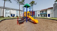 Located at 9430 Concourse Dr in Houston, Texas, this outdoor community space serves as a central gathering and recreational hub within a residential complex. The grounds are anchored by a colorful, multi-feature playground structure equipped with slides, climbing elements, and activity panels, all set on a wood chip safety surface bordered by sturdy edging. Tall pine trees provide natural shade and a sense of enclosure throughout the area. Adjacent to the playground, a covered gazebo with a metal roof offers a shaded seating area with benches, making it an ideal spot for residents to relax and socialize. The open green lawn areas surrounding these amenities are neatly maintained, with trimmed hedges and flowering shrubs adding visual interest along the building perimeters. The two-story residential buildings that frame the courtyard feature white and gray brick exteriors, bold blue entry doors, and balconies with railings on the upper level, giving the complex a cohesive and well-defined architectural character. Walkways connect the various outdoor areas, creating an accessible and functional layout. The overall ambiance is open, community-oriented, and family-friendly, designed to encourage outdoor activity and neighborly interaction.