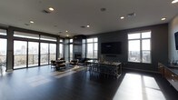 This sophisticated amenity lounge is designed with a modern aesthetic, combining dark tones with generous natural light to create an elegant and inviting atmosphere. The space is anchored by dramatic floor-to-ceiling windows and glass sliding doors that wrap around the room, flooding the interior with daylight and offering expansive panoramic views of the city skyline and surrounding treetops.

Dark hardwood flooring runs throughout, complementing the deep charcoal and slate-toned walls that lend the space a refined, contemporary character. A sleek, black-framed fireplace serves as a striking focal point, providing both warmth and visual interest at the heart of the room.

The lounge is thoughtfully arranged into distinct functional zones. A casual seating area near the windows features plush armchairs, ideal for relaxed conversation or enjoying the views. A dining or collaborative work area offers a long communal table with modern metal chairs, accommodating group gatherings or meetings. A large wall-mounted flat-screen television adds a media and entertainment dimension to the space.

Recessed ceiling lighting provides consistent illumination throughout, while the abundance of natural light from multiple window exposures enhances the open, airy feel of the interior. The overall ambiance is polished and contemporary, suited for both social events and everyday use.