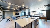 Located at 880 W 200 S in Salt Lake City, Utah, this well-appointed single-floor amenity space offers a thoughtfully designed open-plan layout that maximizes functionality and social interaction. The space centers around a striking oversized kitchen island with a crisp white quartz countertop, an integrated sink, and seating for multiple guests — ideal for casual gatherings and communal use.

The lounge area is generously furnished with plush neutral sofas, modular ottomans, and a large wall-mounted flat-screen TV, creating an inviting media and entertainment hub. A ceiling-mounted projector adds an additional layer of entertainment versatility. The dark, textured ceiling treatment throughout lends an industrial-chic aesthetic, complemented by recessed lighting and exposed track rails.

Large windows line the perimeter walls, flooding the space with natural light and offering open views of the surrounding Salt Lake City neighborhood. A unique roll-up glass garage door opens the space to an outdoor balcony, seamlessly blending interior and exterior living.

A dedicated game area features a foosball table alongside curated wall art, adding a recreational dimension to the space. Light hardwood flooring flows throughout, providing a warm contrast to the deep charcoal and teal accent walls. The overall ambiance is modern, urban, and welcoming — designed to foster community and comfort.