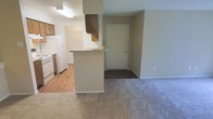 This single-floor unit presents an open-concept layout that connects the kitchen and living area in a practical, efficient design. The kitchen is equipped with white appliances, including a full-size refrigerator and range, and features warm wood-toned cabinetry with overhead fluorescent lighting. A peninsula-style breakfast bar with a laminate countertop serves as a natural divider between the kitchen and the adjacent living space, offering additional prep or dining surface area. The kitchen is finished with hardwood-style flooring, while the living area transitions to wall-to-wall carpeting in a neutral beige tone. A dedicated entryway is positioned near the living area, providing a clear and defined point of entry. The walls throughout are painted in a soft, neutral beige, contributing to a cohesive and understated aesthetic. A doorway visible from the kitchen leads to what appears to be a laundry or utility area, adding to the unit's functional appeal. White baseboards frame each room cleanly. Overall, the space offers a straightforward, low-maintenance interior suited for everyday living.