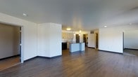 This single-floor space showcases a clean, contemporary design with an open-concept layout that seamlessly connects the living and kitchen areas. Rich, dark hardwood-style flooring runs throughout, providing a warm contrast to the crisp white walls and light gray ceilings. The kitchen is well-appointed with light wood cabinetry, a dark countertop island that doubles as a breakfast bar, a stainless steel refrigerator, and a dishwasher. Pendant lighting hangs above the island, complemented by recessed lighting and a flush-mount ceiling fixture that collectively provide ample illumination. A dedicated in-unit laundry area is conveniently tucked just beyond the kitchen, housing a stacked washer and dryer unit. The open living area is generously proportioned, offering flexible furniture arrangements and a sense of spaciousness. Clean architectural lines, minimal ornamentation, and a neutral color palette throughout lend the space a modern, move-in-ready aesthetic. A hallway visible in the background suggests additional private spaces, completing this functional and well-designed layout.