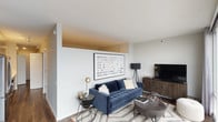 This single-floor residence showcases a cohesive modern aesthetic carried consistently across all rooms. The living area serves as the central gathering space, featuring dark hardwood-style flooring, crisp white walls, and a distinctive architectural soffit detail near the ceiling that adds visual depth. A tufted navy velvet sofa anchors the space, complemented by a patterned area rug, minimalist side tables, and a media console. Large framed artwork above the sofa adds a focal artistic element.

The kitchen is efficiently designed with a clean, contemporary finish. White upper cabinetry contrasts with light wood-toned lower cabinets and bright white countertops. Stainless steel appliances, including a gas range, over-the-range microwave, dishwasher, and French door refrigerator, provide a fully equipped cooking space. A white tile backsplash completes the polished look.

The bedroom offers a calm, restful atmosphere with warm lighting provided by wall-mounted sconces flanking an upholstered channeled headboard. A built-in closet system with shelving and hanging rods provides organized storage.

The bathroom maintains the modern theme with light wood-grain cabinetry, a white undermount sink, clean-lined fixtures, and a large vanity mirror illuminated by a horizontal light bar above.
